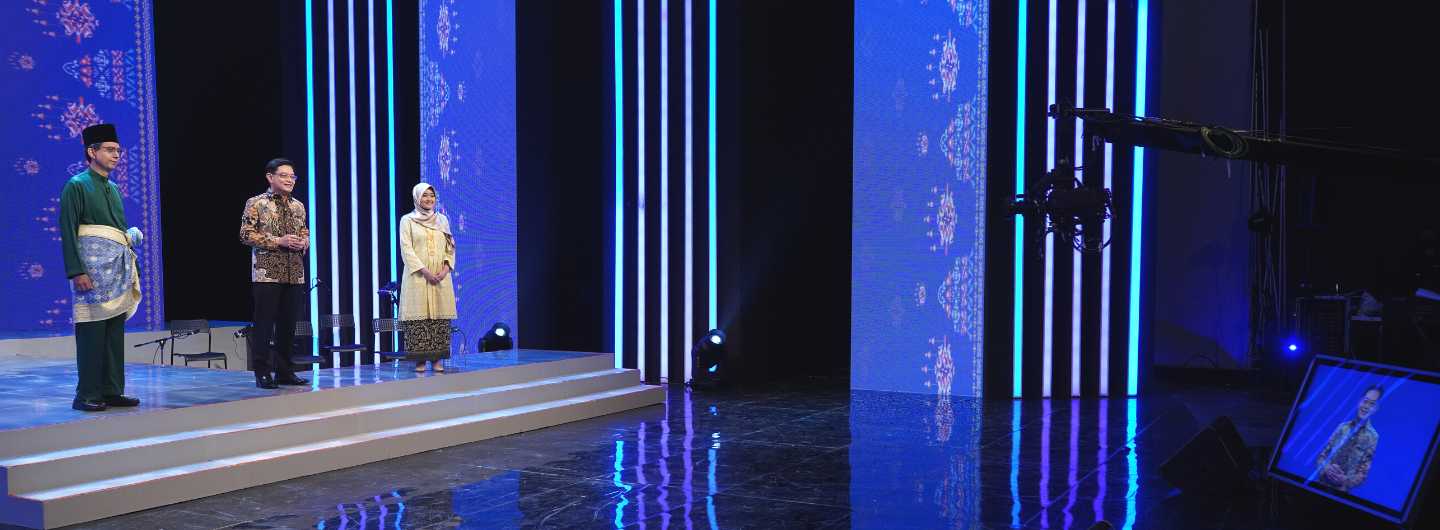 Three people on a stage, blue patterned backdrop, camera equipment visible.